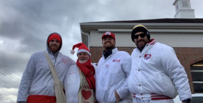 Four people stand outside in front of a brick building, wearing white Charity Newsies jackets, red hats and scarves, and carrying bags for donations.